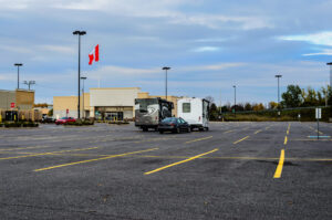 Freshly striped parking lot with yellow lines, a few parked vehicles, and a large commercial building in the background under a cloudy fall sky. A Canadian flag is visible near the center of the image.