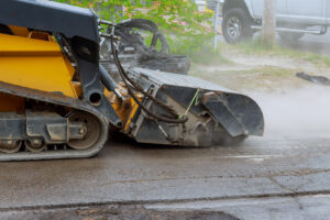 Close-up of an asphalt milling machine grinding a road surface, with visible dust and hydraulic hoses, during pavement repair or resurfacing work.