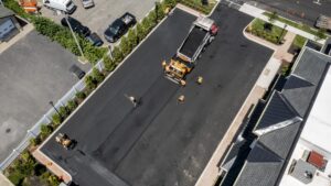 Aerial view of an asphalt paving crew resurfacing a commercial parking lot. Several workers in orange safety gear operate paving and compacting equipment around a dump truck unloading hot asphalt, surrounded by landscaped edges and adjacent buildings.