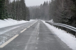 A snowy two-lane rural road surrounded by pine trees, with light snow covering the asphalt and a single car in the distance under overcast skies.