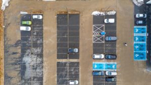 Aerial view of a commercial parking lot in winter showing snow piles, faded asphalt, marked parking spaces, and accessible parking areas.
