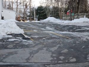 Parking lot with snow piles and visible asphalt surface cracking and ice damage caused by freeze-thaw conditions in winter.