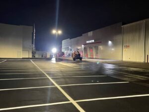 Freshly painted parking lot lines outside a commercial service center at night, with a striping machine in use under bright overhead lights.