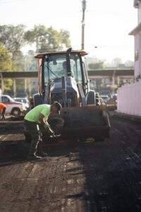 Worker performing asphalt milling and patching on a roadway, using a shovel in front of a loader during early morning light.